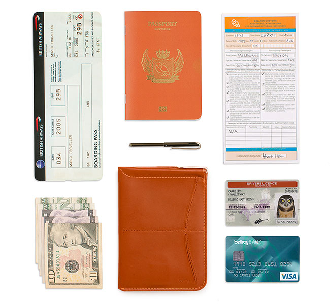 Passport Sleeve Tanの周りにクレジットカード2枚、出入国カード、パスポート、マイクロペン、航空券、二つ折り紙幣5枚を並べた画像