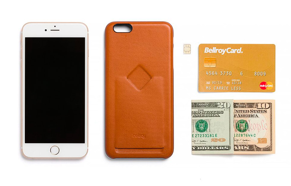 iPhoneケースの隣に置かれたベルロイのフォンケース1カードキャラメルチャコールの正面画像とクレジットカード1枚、折り畳んだ紙幣2枚、SIMカードの写真を収納できる貴重品の例として並べた様子。